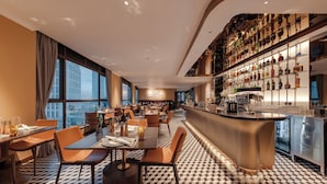 A modern restaurant with a checkered floor, a bar area with shelves of bottles, and a city view through large windows.