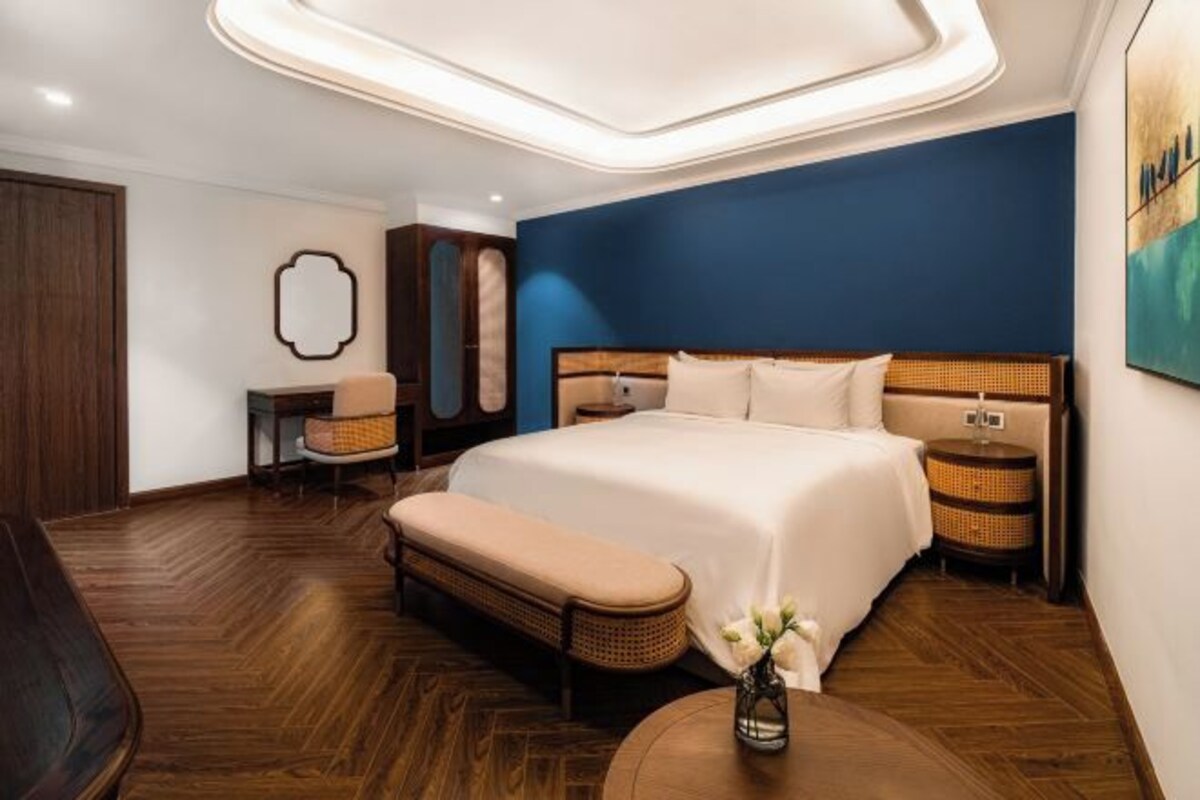 A modern hotel room with a large bed, a bench, a small table, and a wall-mounted TV.