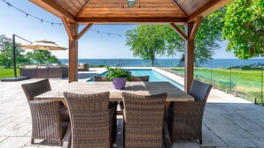 A covered patio with a pool, wicker furniture, and a view of the sea. - Lakefront Luxury | Pool, Hot Tub, Wine Route (Kingsville)