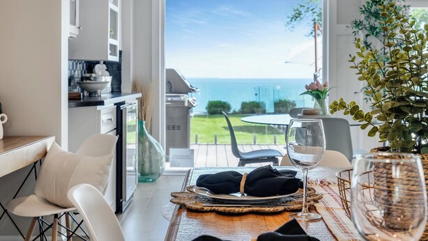 A dining area with a view of the sea, a wooden table set with plates, glasses, and a vase. - Lakefront Luxury | Pool, Hot Tub, Wine Route (Kingsville)