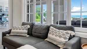 A grey sofa with a white throw blanket and patterned pillows, a large window with white frames, and a view of the outdoors. - Lakefront Luxury | Pool, Hot Tub, Wine Route (Kingsville)