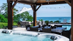 A hot tub with a wooden exterior, a covered seating area with outdoor furniture, and a view of the ocean. - Lakefront Luxury | Pool, Hot Tub, Wine Route (Kingsville)