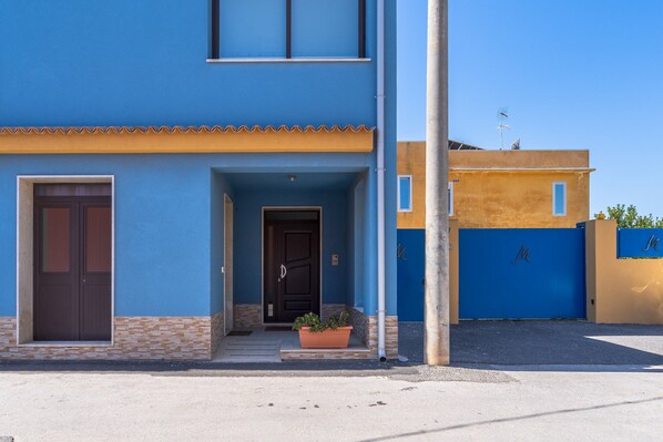 Un bâtiment à deux étages avec une façade bleue, une porte marron et une petite entrée agrémentée d’un pot de fleurs.