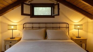 Un dormitorio con techo inclinado, una cama con ropa de cama blanca, dos mesitas de noche con lámparas y una ventana sobre la cama.