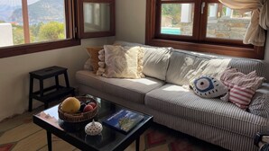 Una sala de estar acogedora con un sofá a rayas, una mesita con fruteros y un libro, y ventanas amplias con vistas a la vegetación y montañas.