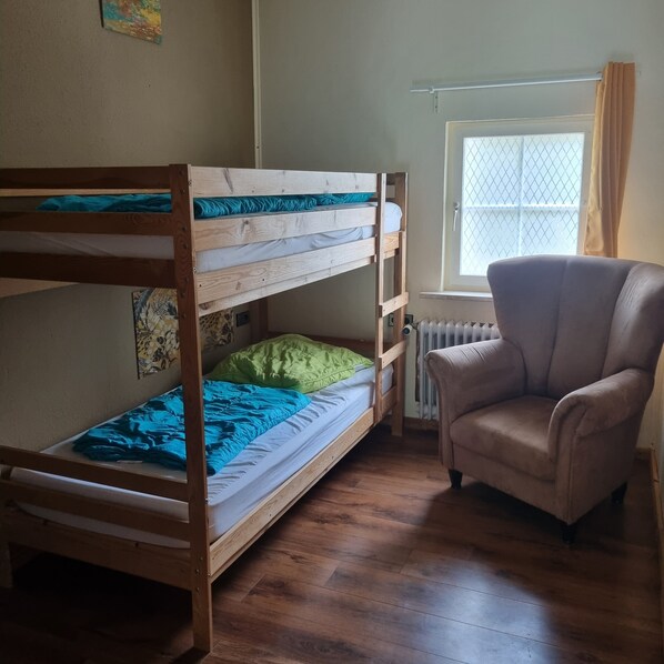 A bunk bed with a green pillow and blue blanket, a beige armchair, and a window with a radiator.