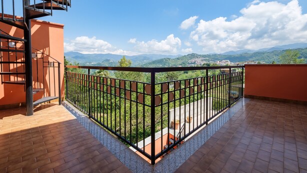 Un balcone con ringhiera in metallo, pavimento piastrellato e vista su colline e montagne verdi.