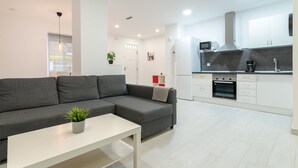 Un salon moderne avec un canapé gris, une table basse blanche et une plante en pot. L’espace cuisine comprend des meubles blancs, un four à micro-ondes, une cuisinière et une machine à café.