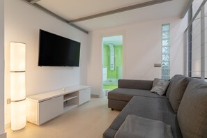 Ein modernes Wohnzimmer mit einem Flachbildfernseher, einer weißen Unterhaltungseinheit, einem grauen Sofa und einer Glastür zu einem hellen Raum.