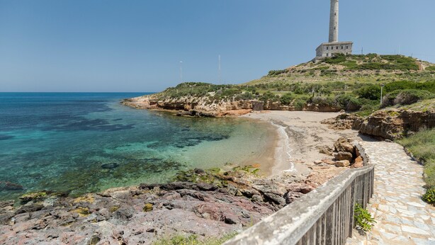 A coastal landscape with a lighthouse, clear blue sea, rocky shore, and a stone pathway.