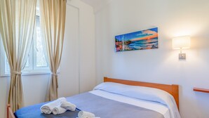 A neatly made bed with white and blue bedding, a painting of a sunset on the wall, and a lamp on the nightstand. - Vacation Home "San Lorenzo" with Private Terrace, Private Garden and A/C (San Lorenzo)