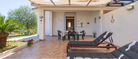 A covered outdoor area with lounge chairs, a table with chairs, and a view of the garden.