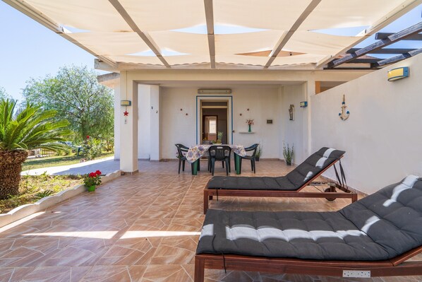 A covered outdoor area with lounge chairs, a table with chairs, and a view of the garden.