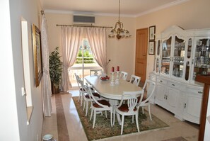 Uma sala de jantar com uma mesa branca, cadeiras, um lustre e um aparador com portas de vidro.