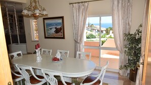 Un espace repas avec une table ronde, des chaises blanches, un lustre, un tableau et une vue sur la mer.