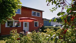 Una casa de dos pisos con fachada roja, un porche cubierto con un paraguas rojo y un jardín con diversas plantas y un árbol que da frutos rojos.