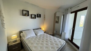 A bedroom with a bed, bedside lamps, a small table, a radiator, and a view through a window. - Saint-Cyprien: Renovated T2 cabin for 6, pool, parking, 100m from the beach (Saint-Cyprien)