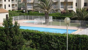 A swimming pool surrounded by a white fence, palm trees, and multi-story buildings with balconies. - Saint-Cyprien: Renovated T2 cabin for 6, pool, parking, 100m from the beach (Saint-Cyprien)