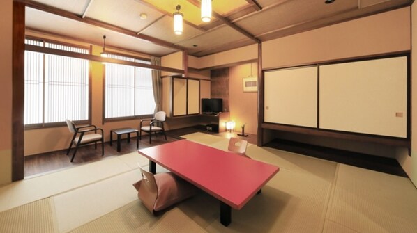 Ein traditioneller japanischer Raum mit Tatami-Bodenbelag, einem niedrigen Tisch, Stühlen, einem Fernseher und Schiebetüren.