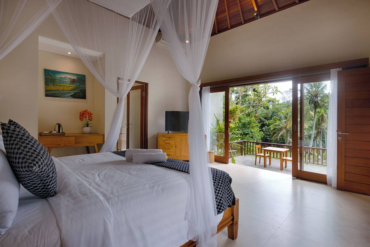 A bedroom with a bed, a desk, a television, and a balcony with a view of the outdoors.