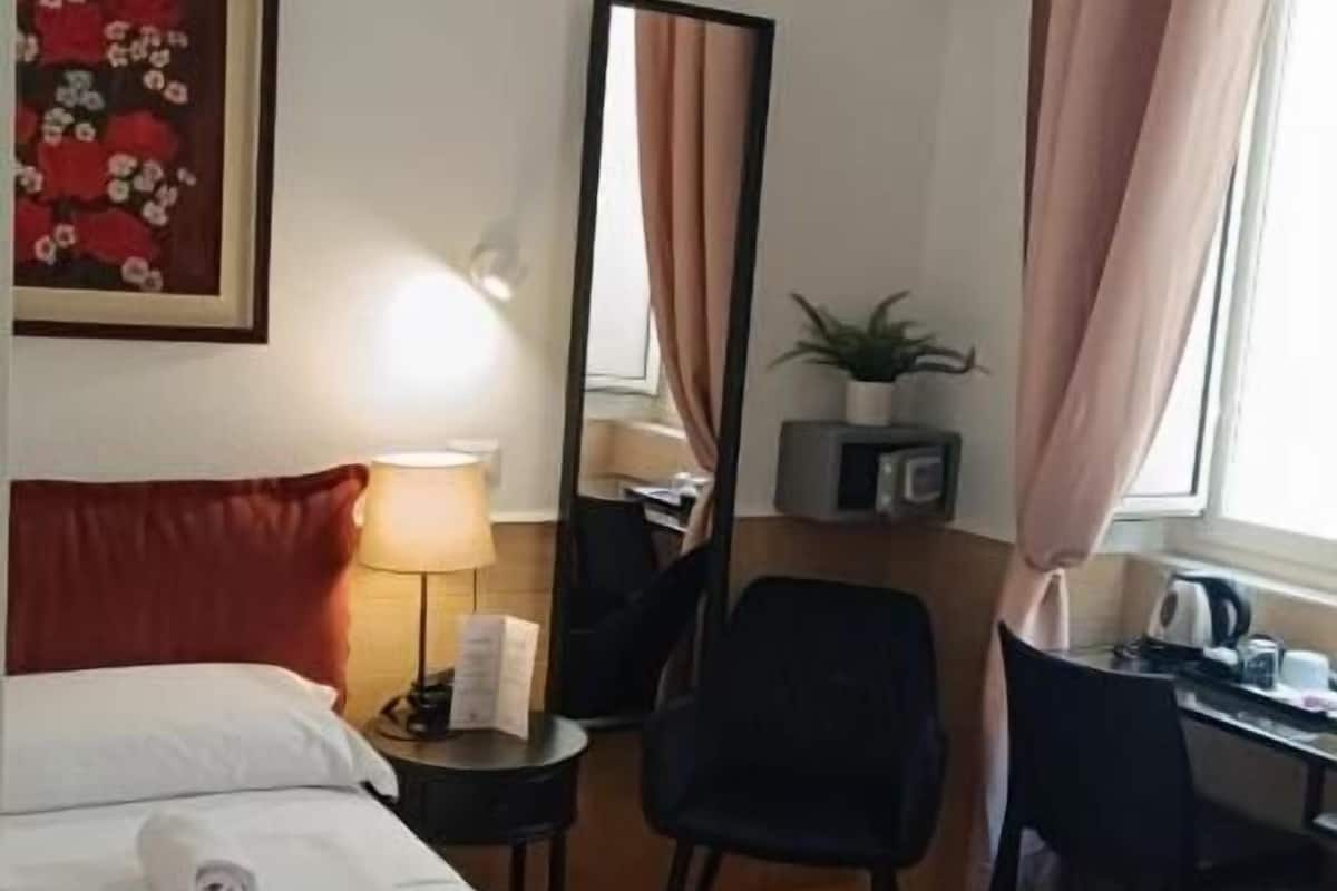 A hotel room with a bed, a desk, a chair, a mirror, a lamp, and a window with curtains.