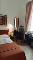 Una camera d'albergo con un letto, una scrivania, una sedia, uno specchio, una lampada e una finestra con tende.