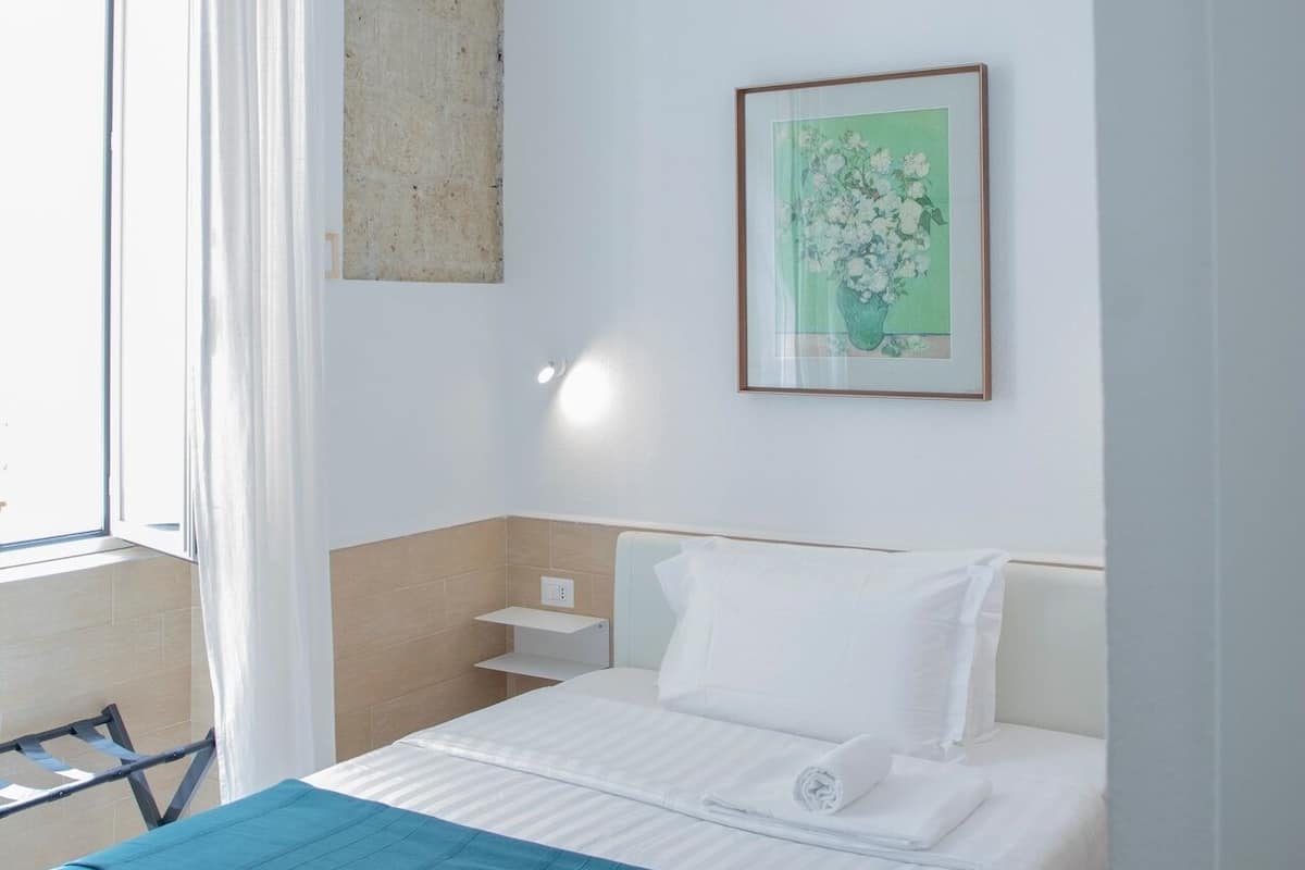 A hotel room with a bed, a nightstand, a framed floral artwork, and a window with curtains.