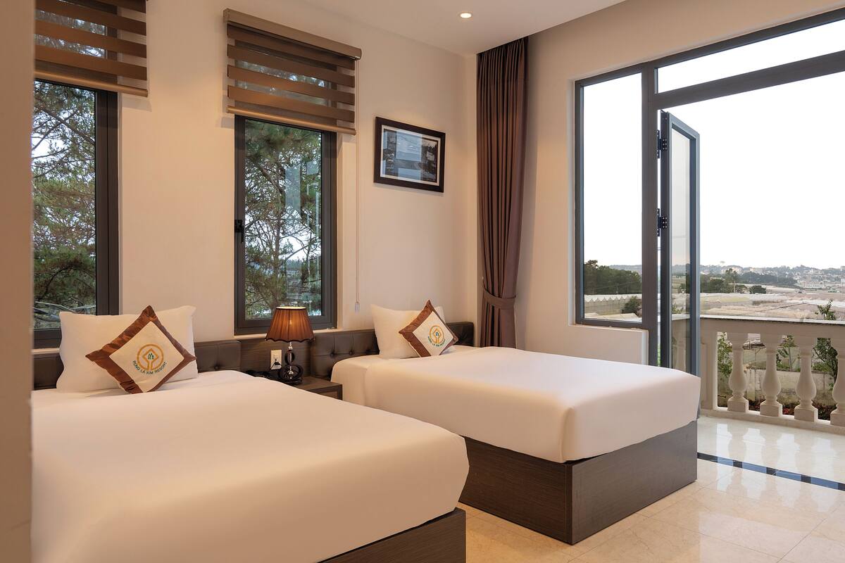 Quarto de hotel com duas camas, uma janela com persianas, uma luminária e vista para a paisagem.