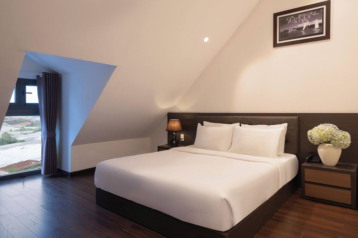 Quarto moderno com uma cama grande, mesa de cabeceira, abajur, quadro na parede e janela com cortinas.
