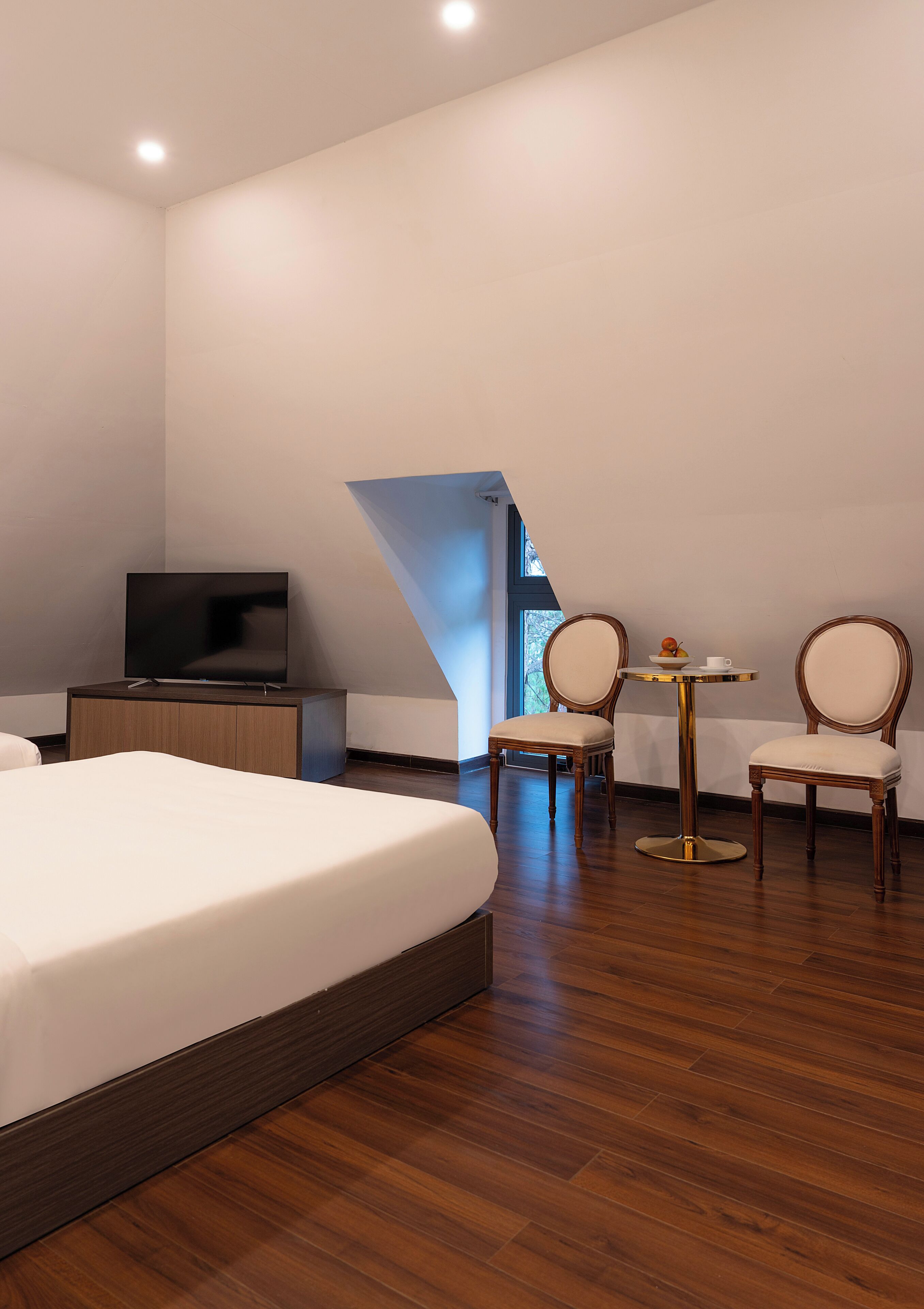 Una habitación de hotel moderna con una cama grande, un televisor sobre un mueble de madera, dos sillas alrededor de una mesita y vistas a un cielo azul a través de una ventana.