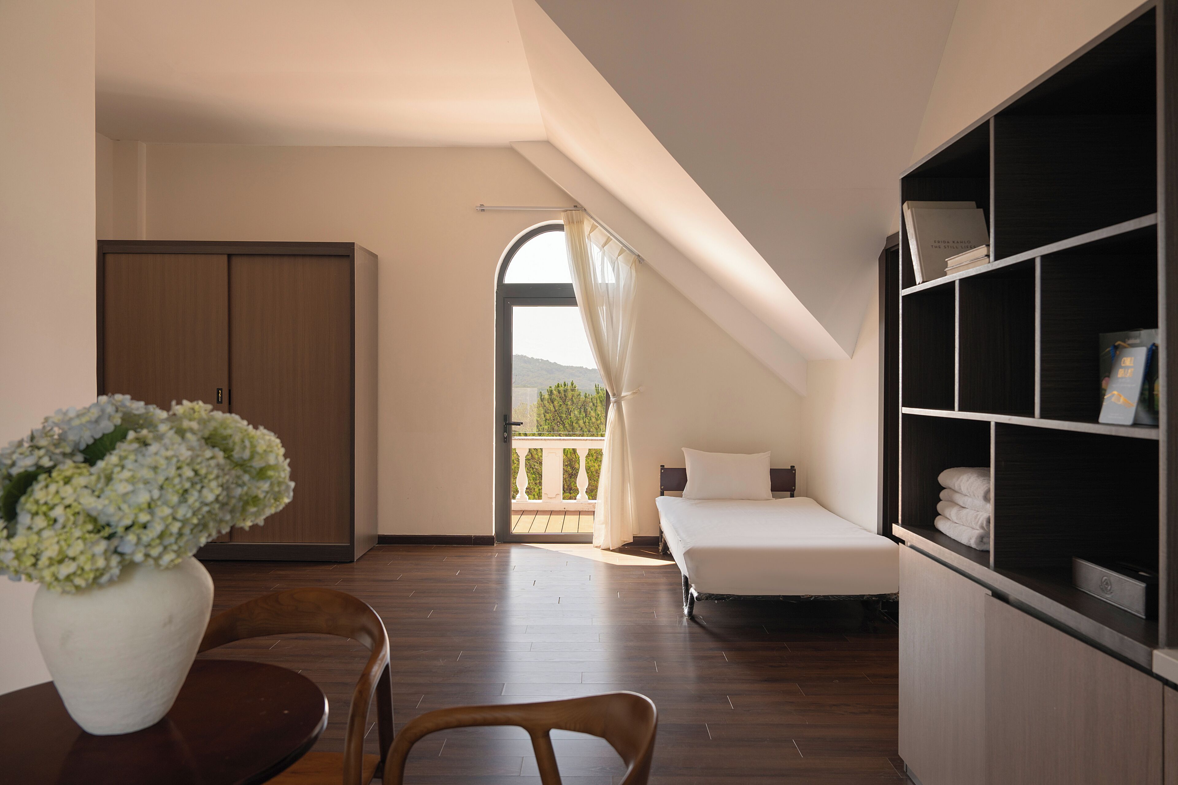 A bedroom with a bed, a wardrobe, a desk with a chair, a bookshelf, and a view of a balcony.