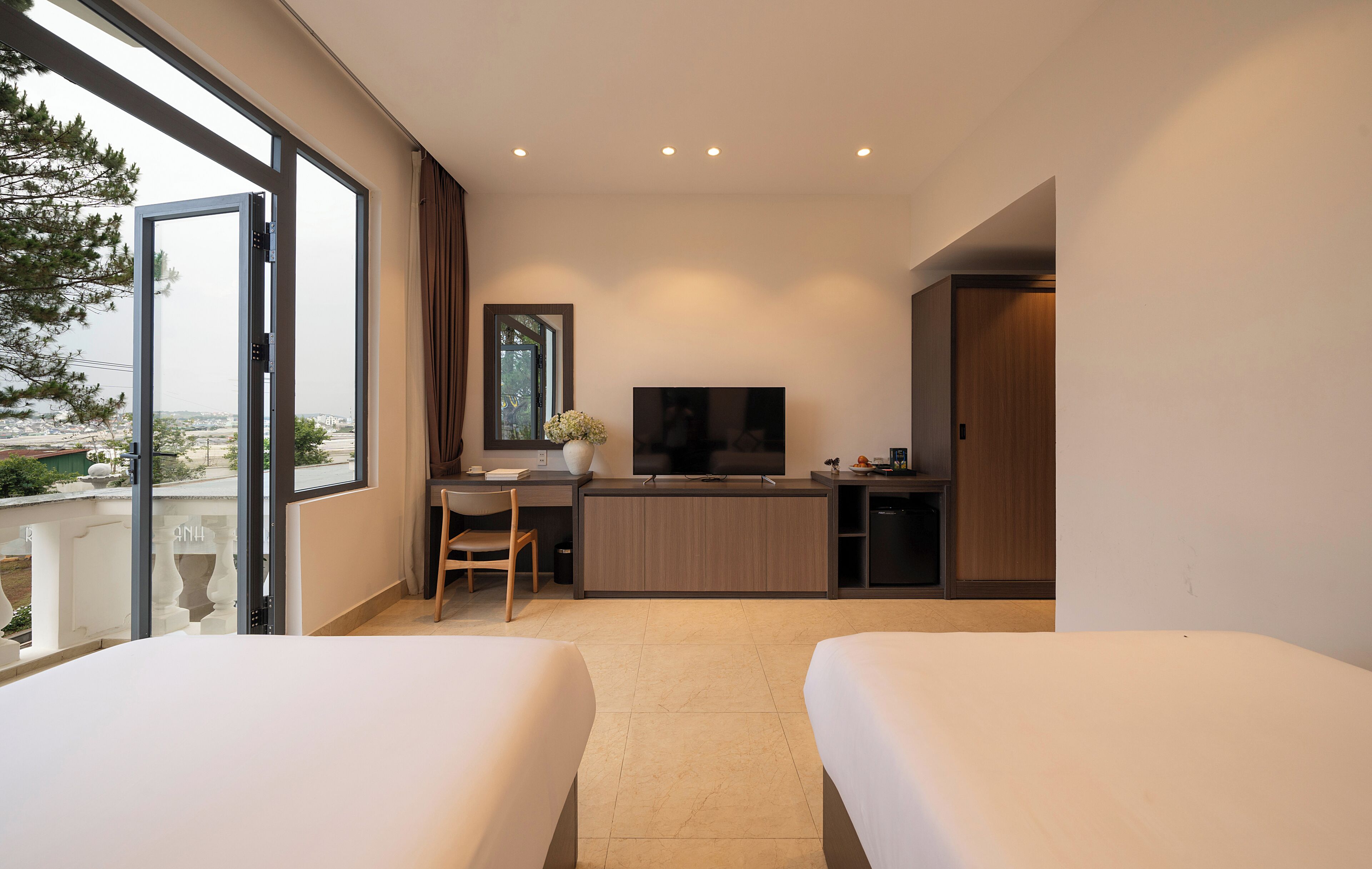 A hotel room with two beds, a desk with a chair, a TV, and a balcony with a view.