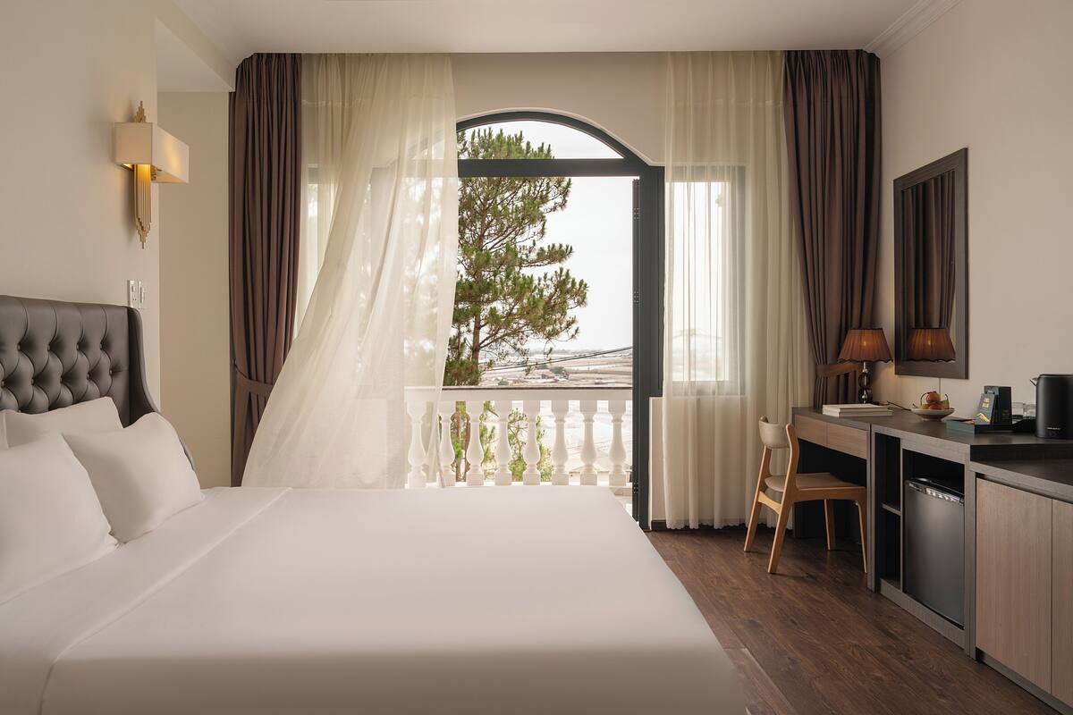 Quarto de hotel com cama, escrivaninha, cadeira, abajur e vista pela janela.