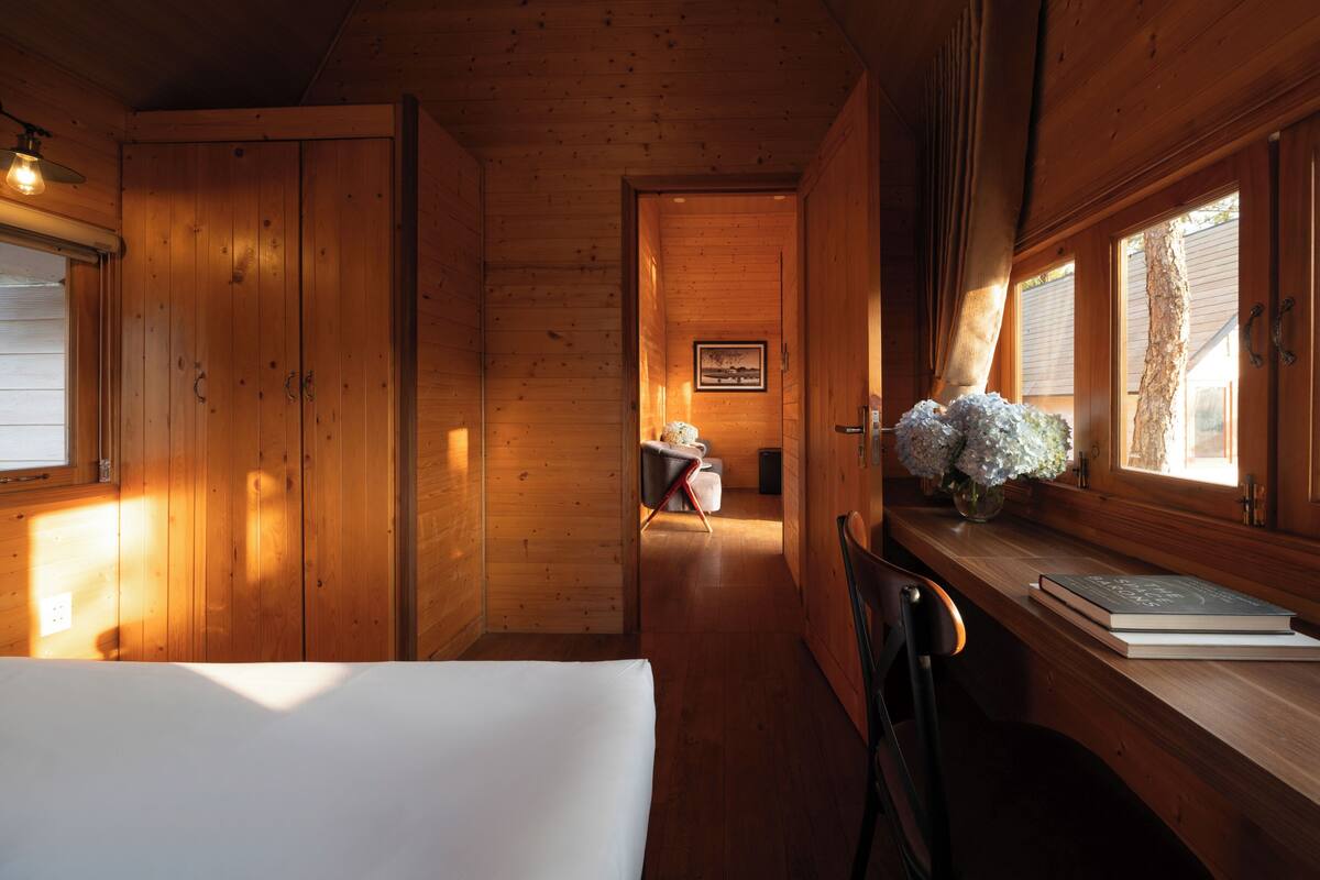 Interior de uma cabana de madeira com uma cama, uma escrivaninha com livros, uma cadeira e uma janela com vista para o exterior.