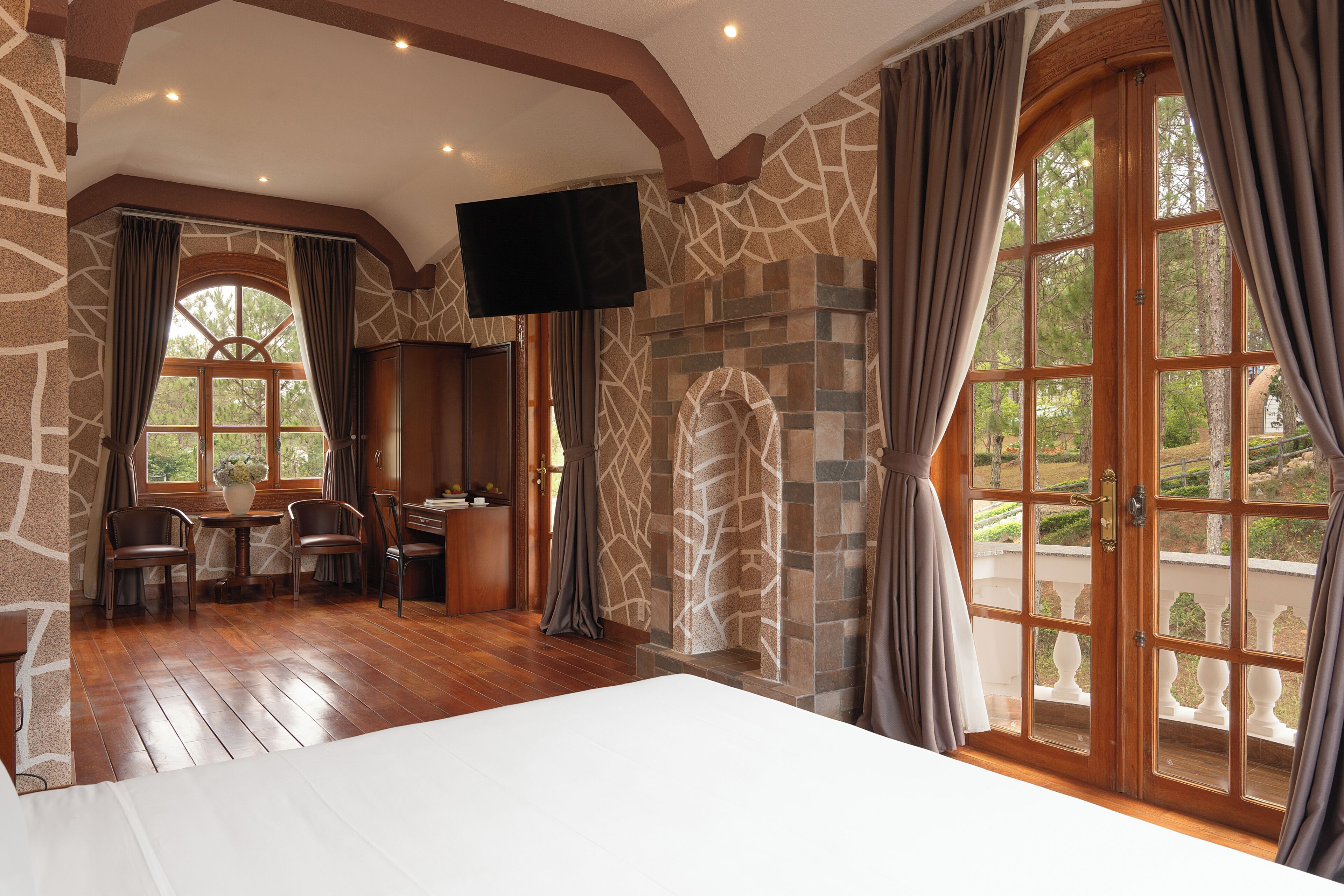 Una habitación con una cama grande, paredes de piedra, chimenea, televisor y balcón con vistas a árboles.
