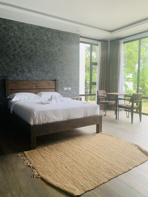 A modern bedroom with a large bed, a wooden headboard, a small table with chairs, and a view of greenery through large windows.