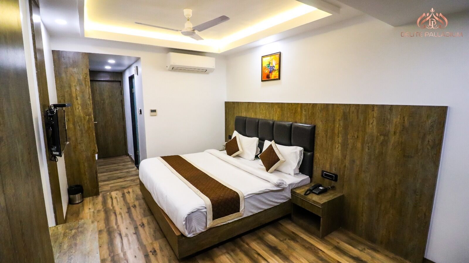 Una habitación de hotel moderna con suelo de madera, una cama grande con cabecero, una mesita de noche con teléfono y un equipo de aire acondicionado en la pared.