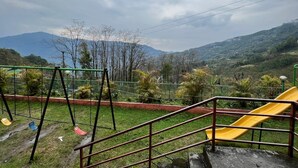 Un parc infantil amb cadira de balanç i tobogan, envoltat de vegetació i turons.