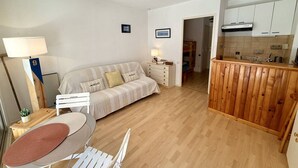 Ein kompakter Wohnbereich mit Sofa, Esstisch, Küchenzeile und einem Hochbett im Hintergrund.