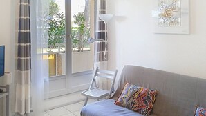 A living room with a grey sofa, colorful pillows, a white lamp, and a view of greenery outside. - Charming 1BR Apartment, Garden View, Near Beach & Shops, Parking Available (Bormes-les-Mimosas)