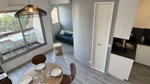 A modern kitchen with white cabinets, a dining area with a round table set for four, and a balcony with a view of greenery. - Cabourg: Renovated Duplex 2P, Direct Beach, Fiber (Cabourg)