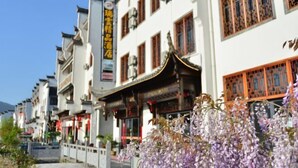 A row of traditional buildings with a signboard in a foreign language, a traditional roof, and a purple flowering plant in the foreground.