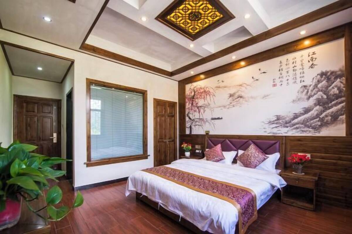 A bedroom with a large bed, wooden flooring, a window with blinds, a wall-mounted artwork, and a ceiling with recessed lighting.