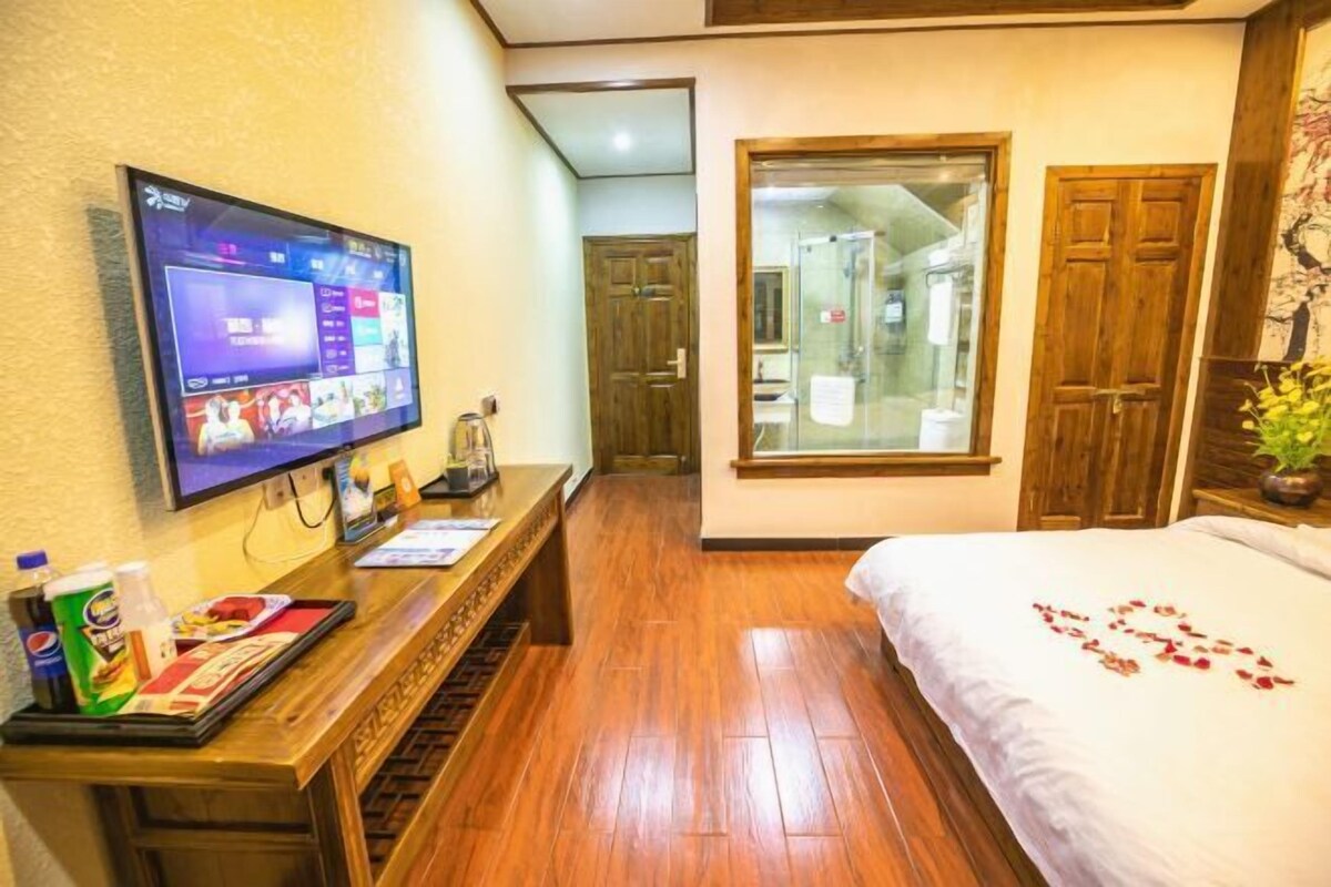 A hotel room with a wooden floor, a bed with white bedding, a TV on the wall, a wooden desk with a tray of items, and a window with a view of a bathroom.