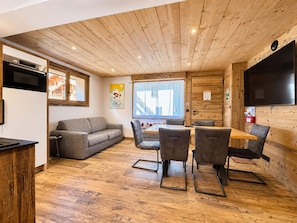 Een gezellige kamer met houten plafond, een bank, een eettafel met stoelen, en een televisie die aan de muur is bevestigd.