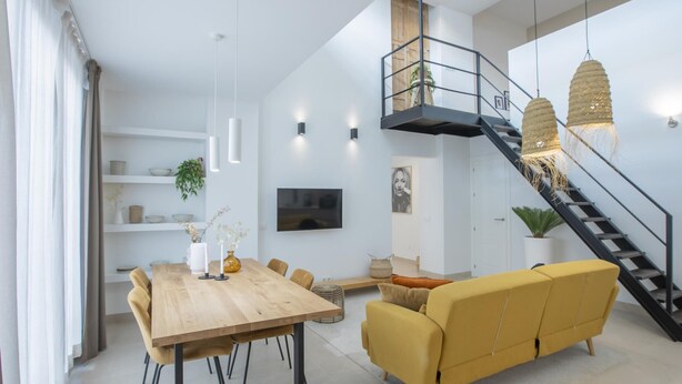 A modern living room with a wooden dining table, yellow sofa, and a staircase with a black railing.