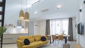 A modern living room with a yellow sofa, dining area, kitchen, and a TV mounted on the wall.