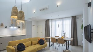 A modern living room with a yellow sofa, a dining area with a table and chairs, and a kitchen with white cabinets and pendant lights.