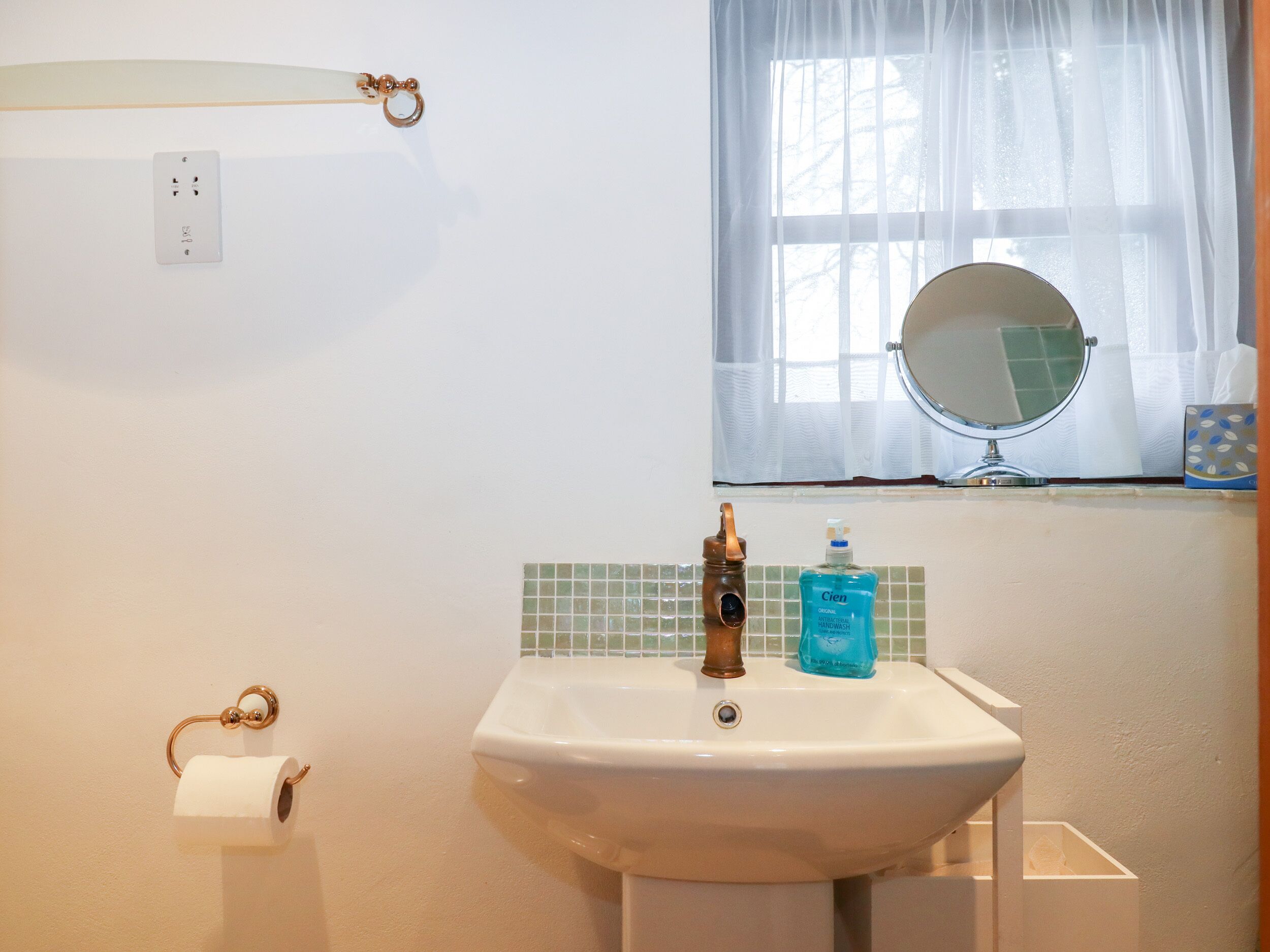 Une salle de bain équipée d’un lavabo, d’un miroir, d’un porte-rouleau de papier toilette et d’une fenêtre avec un rideau.