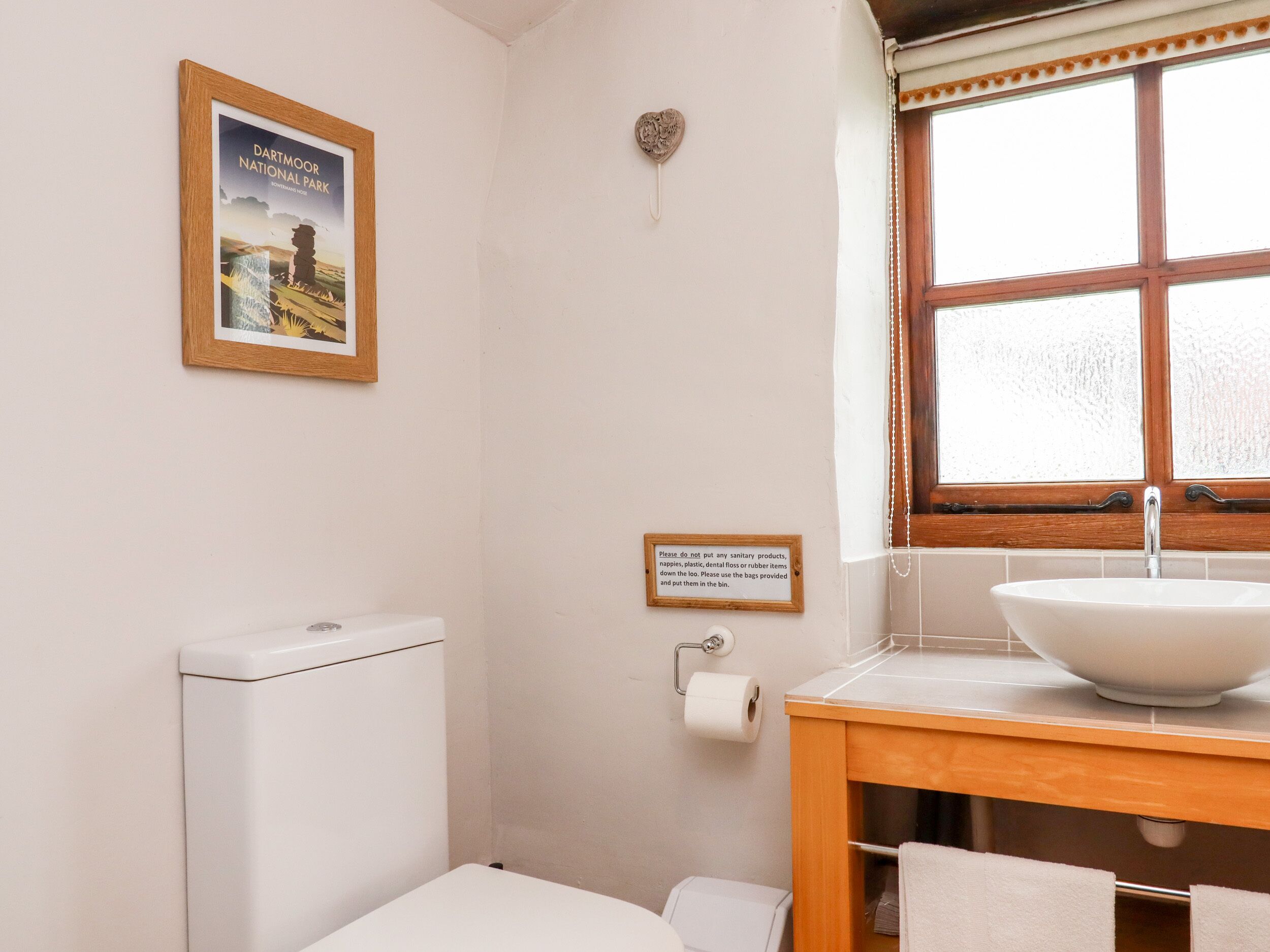 A bathroom with a toilet, a sink, a window, a framed picture, and a sign.